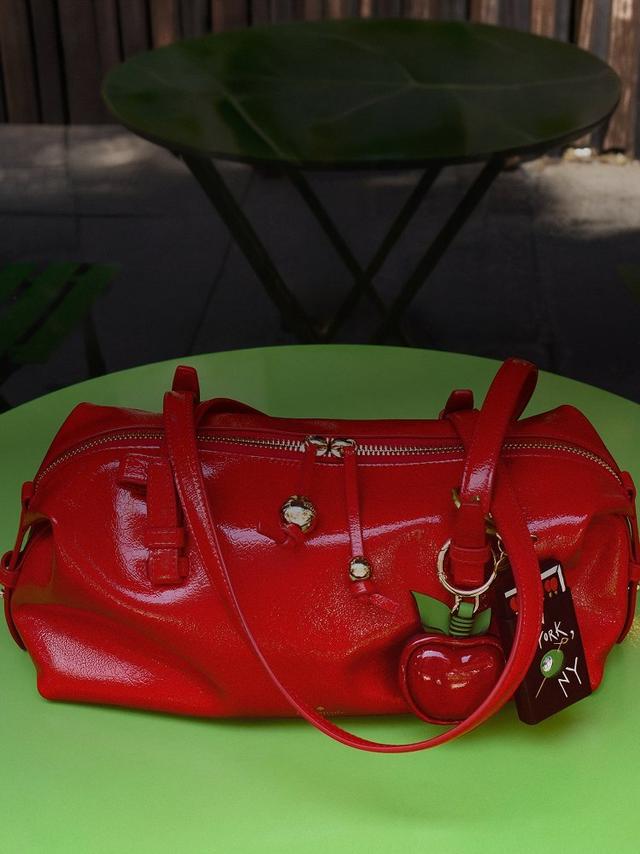 Red leather handbag with dual handles, top zipper, and heart-shaped charm keychain on green surface.