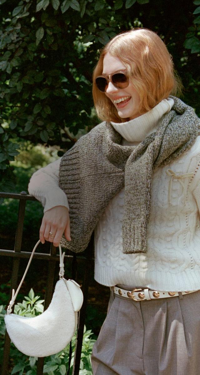 Two women chatting outdoors, one in a brown leather jacket and one in a cream sweater with sunglasses.