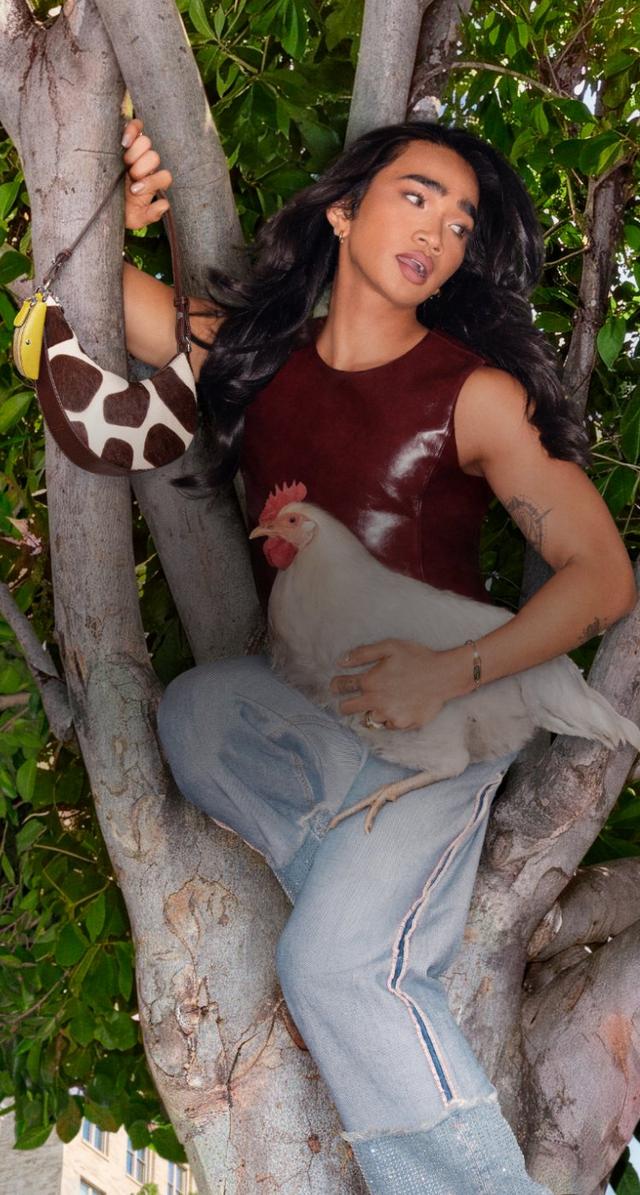 Model wearing a burgundy sleeveless top and holding a giraffe-print handbag, posing with a chicken in a tree.