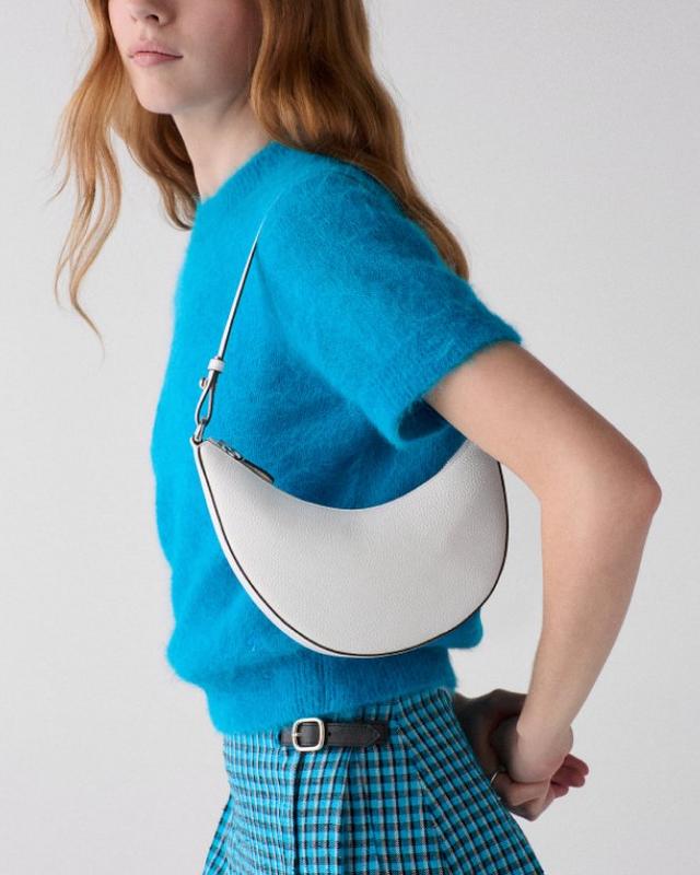 Woman wearing bright blue sweater and plaid skirt carrying white crescent-shaped shoulder bag with black trim.