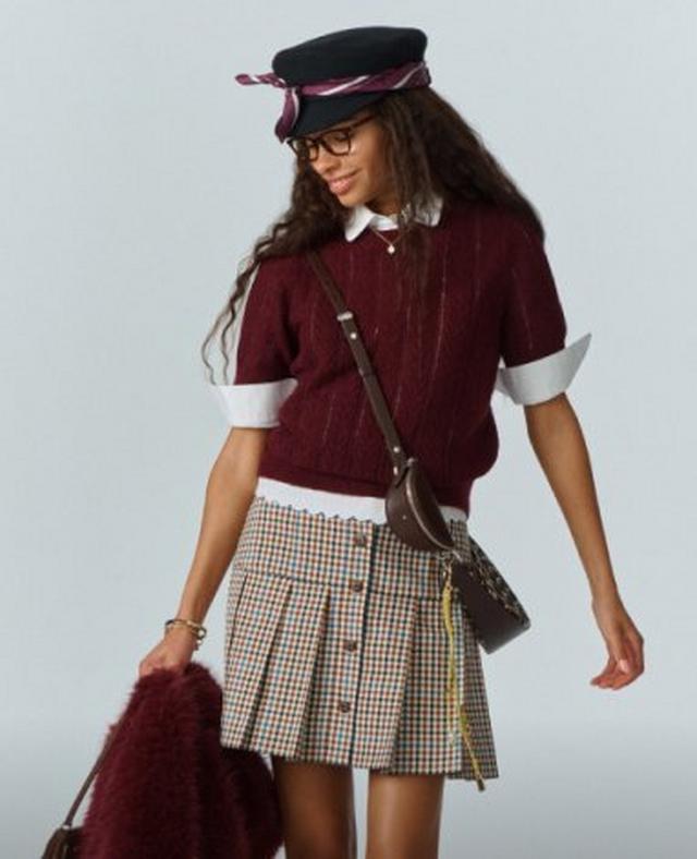 Woman wearing burgundy cable knit sweater, plaid skirt, black cap, and glasses while holding a jacket.