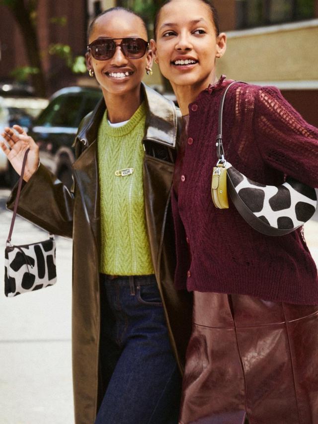 Two smiling women walking together on a city street, carrying matching cow-print handbags.