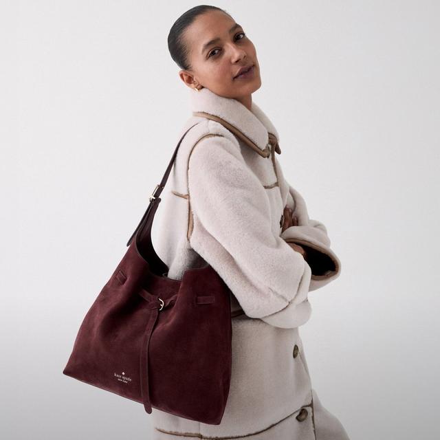 Model wearing cream coat with brown trim carrying a burgundy Kate Spade suede bucket bag on shoulder.