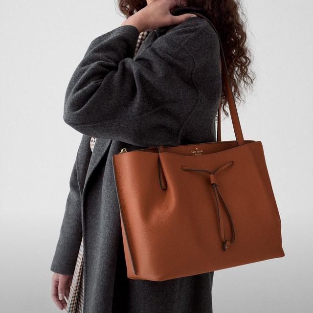 Woman wearing a dark gray coat carrying a brown leather Kate Spade tote bag on her shoulder.