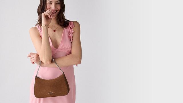 Woman wearing pink dress holding the Kate Spade Margot suede shoulder bag in hot fudge colourway