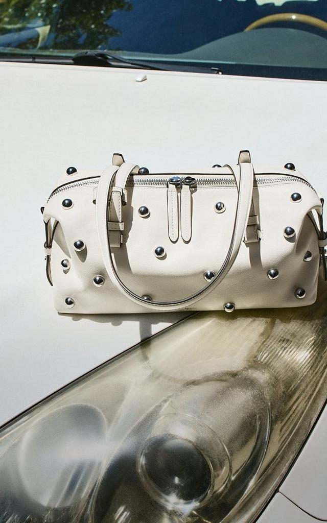 Cream leather handbag with silver metal studs and dual zipper compartments resting on a wooden surface.