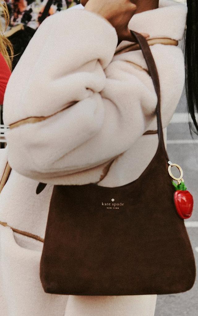Kate Spade brown shoulder bag with red strawberry charm and red dog leash held by person in beige sweater.