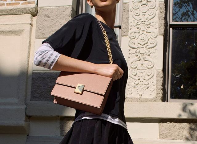 A person in a black and white outfit holds a pink handbag with a gold chain strap, standing against an ornate stone wall with a window. The scene feels chic and stylish.