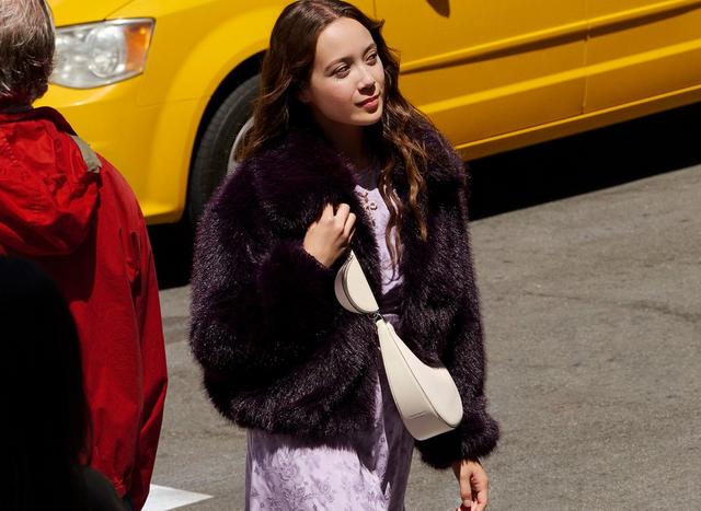 A person in a dark fluffy coat and lavender dress walks on a sunny street beside a yellow vehicle, exuding confidence and style.
