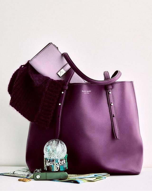 A stylish purple tote bag with a small matching pouch and a dark knit scarf peeking out. Nearby are a New York snow globe, maps, and a green keychain.