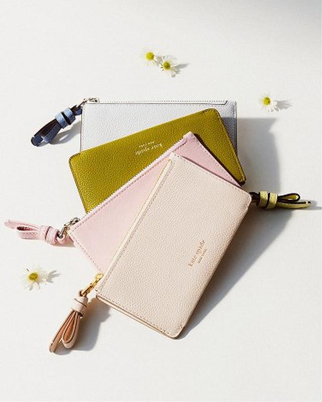 Four pastel-colored wristlet pouches are stacked, featuring shades of pink, olive, and white. Small daisies are scattered around, adding a fresh touch.