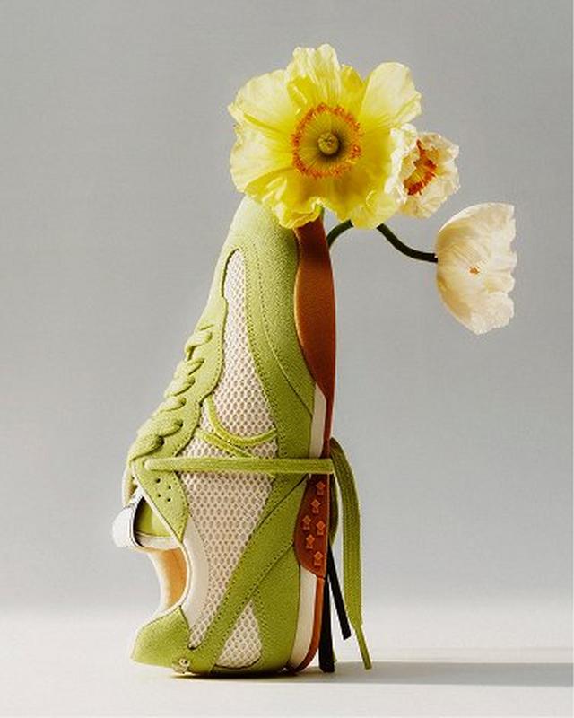A vibrant sneaker with green and white mesh stands on its heel against a neutral background. Yellow flowers emerge from the shoe, creating a fresh, whimsical vibe.