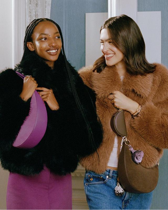 Two women smiling warmly at each other. One in a black fur coat with a purple bag, the other in a brown fur coat with a brown bag, in a cozy, elegant room.