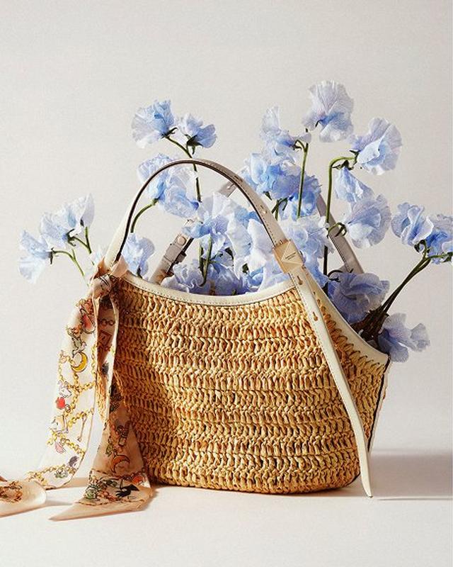 A woven straw handbag with a floral scarf tied to the handle holds delicate pale blue flowers, set against a soft beige background. Elegant and serene.