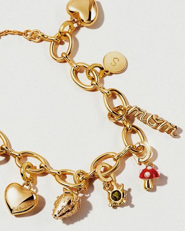 Gold charm bracelet featuring the word "mom," heart, mushroom, shell, and turtle charms. The design is elegant and playful against a white background.